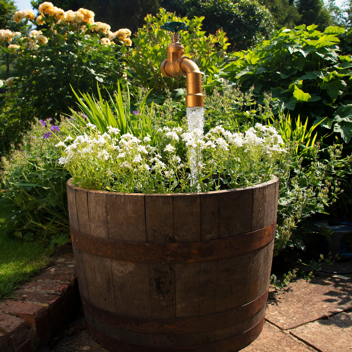 A half oak barrel water feature with a large brass tap installed in the center, surrounded by plants and set in an outdoor setting.