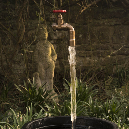 A floating tap water feature with a red gate valve tap attached to a black container, with water flowing into the container, set in a garden environment.
