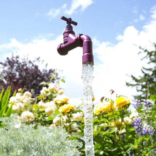 A floating tap water feature with a solid brass tap and soldered copper spout, running water, set in a garden environment with flowers in the background.