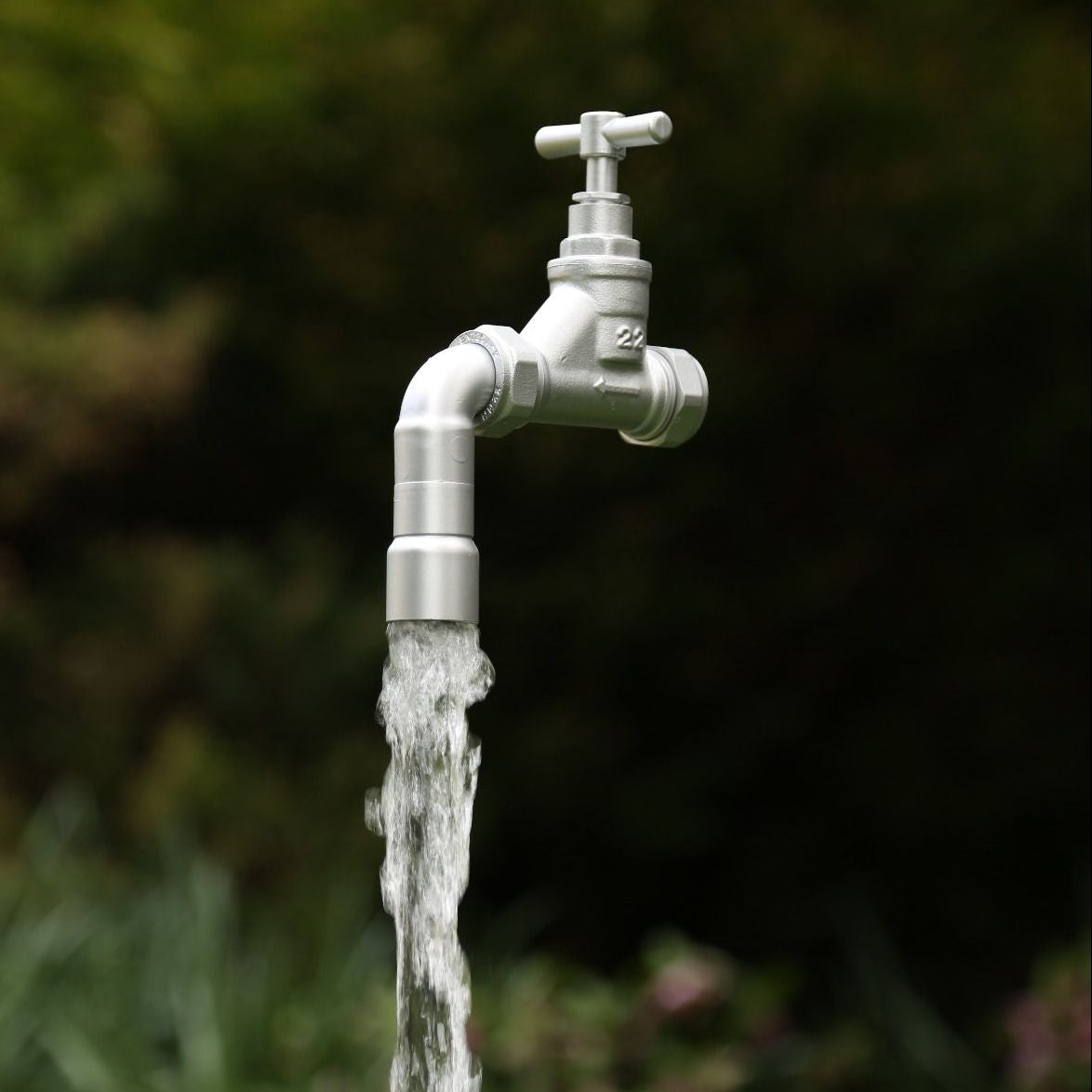 A floating tap water feature with a solid brass tap and soldered copper spout, appearing to float from the ground, with real running water, set in a garden environment with flowers in the background.
