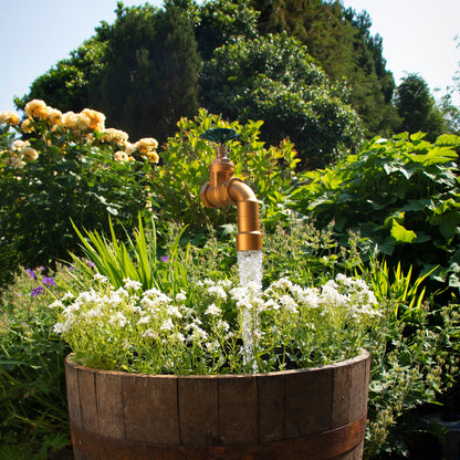 A large floating tap installed on top of a barrel surrounded by plants, with a clear sky and trees in the background.