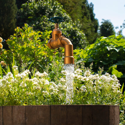 A large floating tap installed on top of a barrel surrounded by plants, with a clear sky and trees in the background.