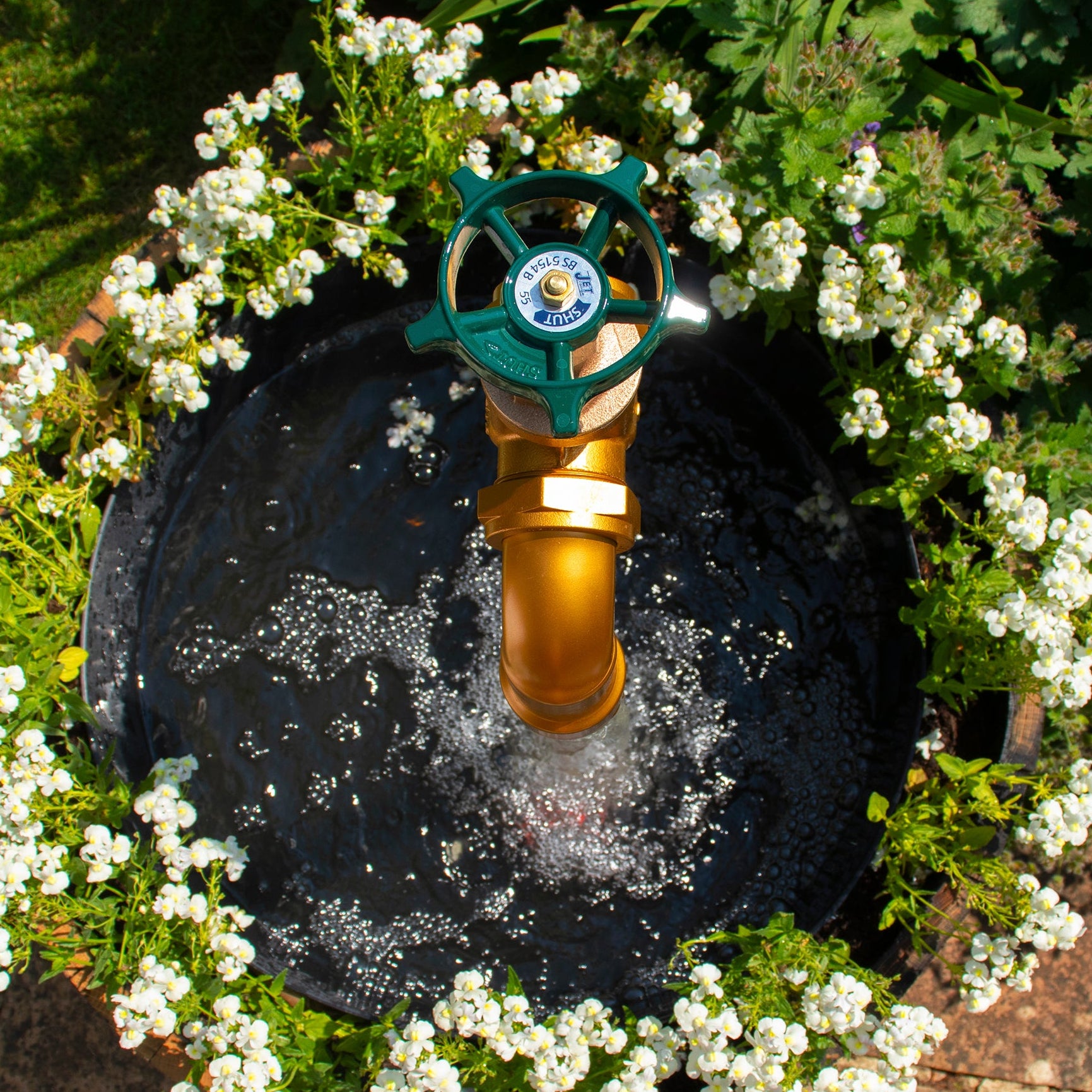 A large floating tap installed on top of a barrel surrounded by plants, with a clear sky and trees in the background.